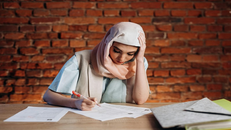 Person sitting at a table with papers, holding a red pen, in front of a brick wall.